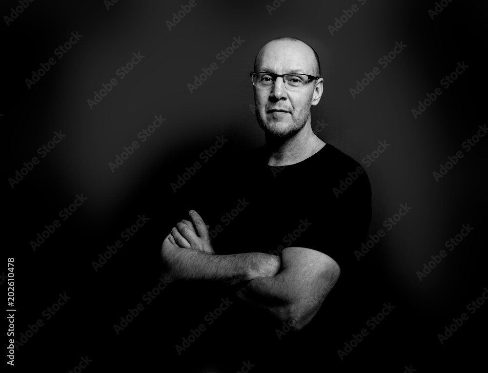 Black and white studio portrait of a middle aged man wearing glasses and smiling with arms crossed in a black t-shirt against a black background