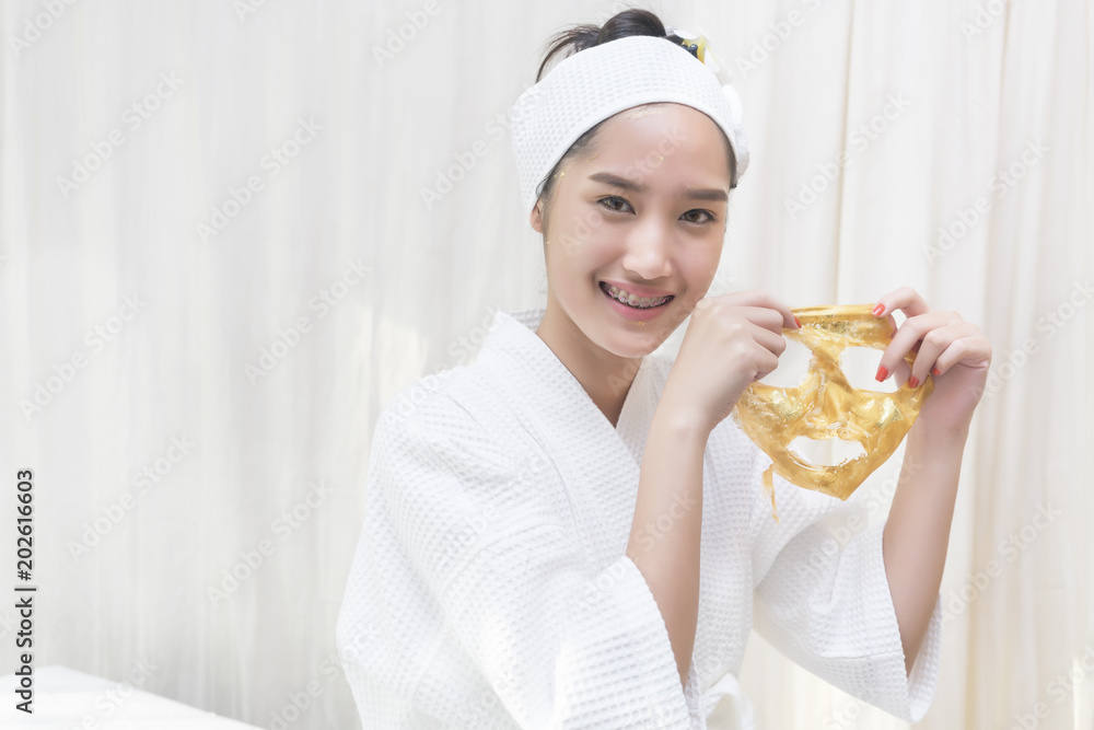 Beautiful young Asian woman in white bathrobe peeling high quality and authentic pure gold facial mask for spa treatment which make her skin glowing, hydrated, and radiant