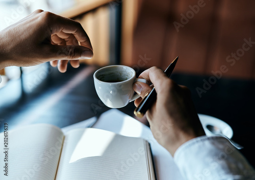 man holds a cup with a drink for writing in his hand and an open notebook