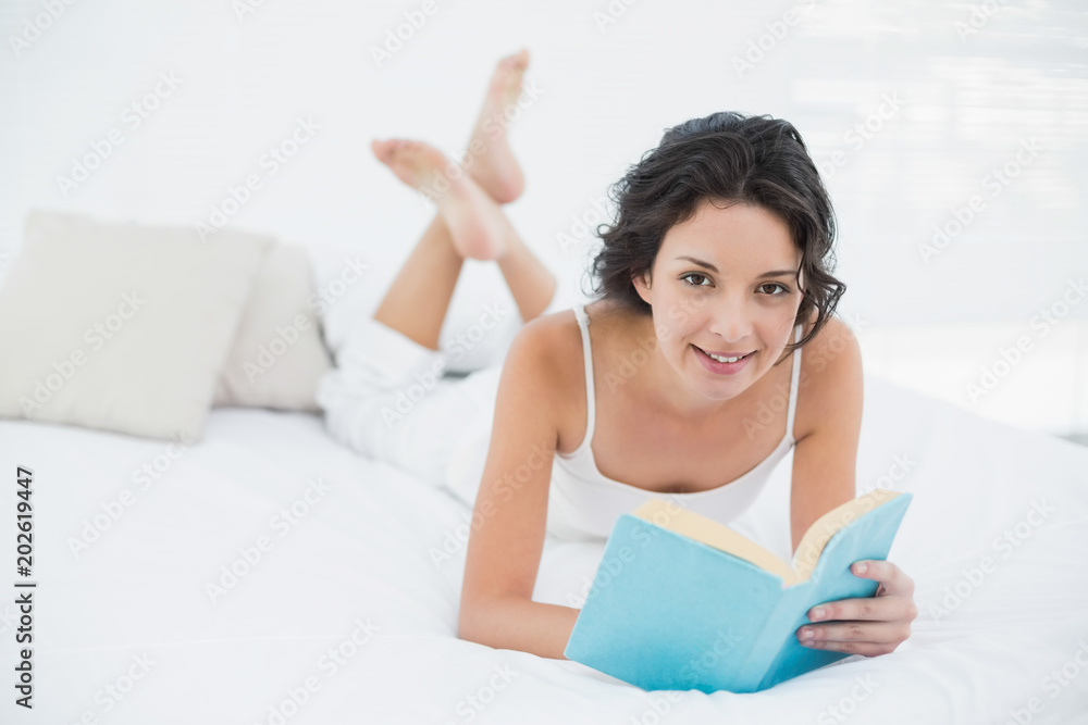 Content casual brunette in white pajamas relaxing on a bed reading a book Content casual brunette in white pajamas relaxing on a bed reading a book