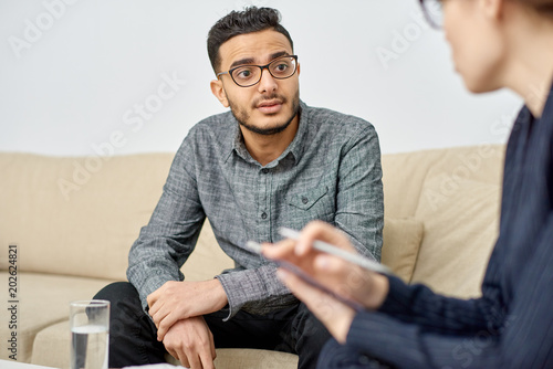 Fototapeta Naklejka Na Ścianę i Meble -  Handsome young man suffering from depression sitting on cozy sofa and listening to highly professional psychologist while she giving him piece of advice