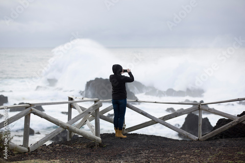 Wild, stormy weather, weavy ocean, rocky shore, wooden fence, woman in black look at the sea. photo