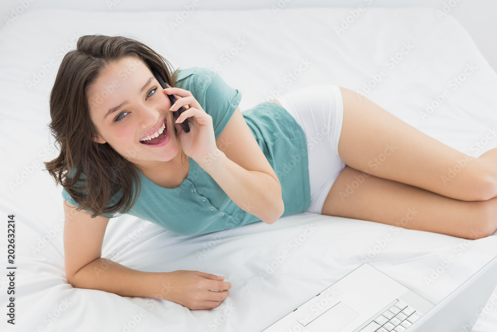 Cheerful casual woman using mobile phone by laptop in bed Cheerful casual woman using mobile phone by laptop in bed