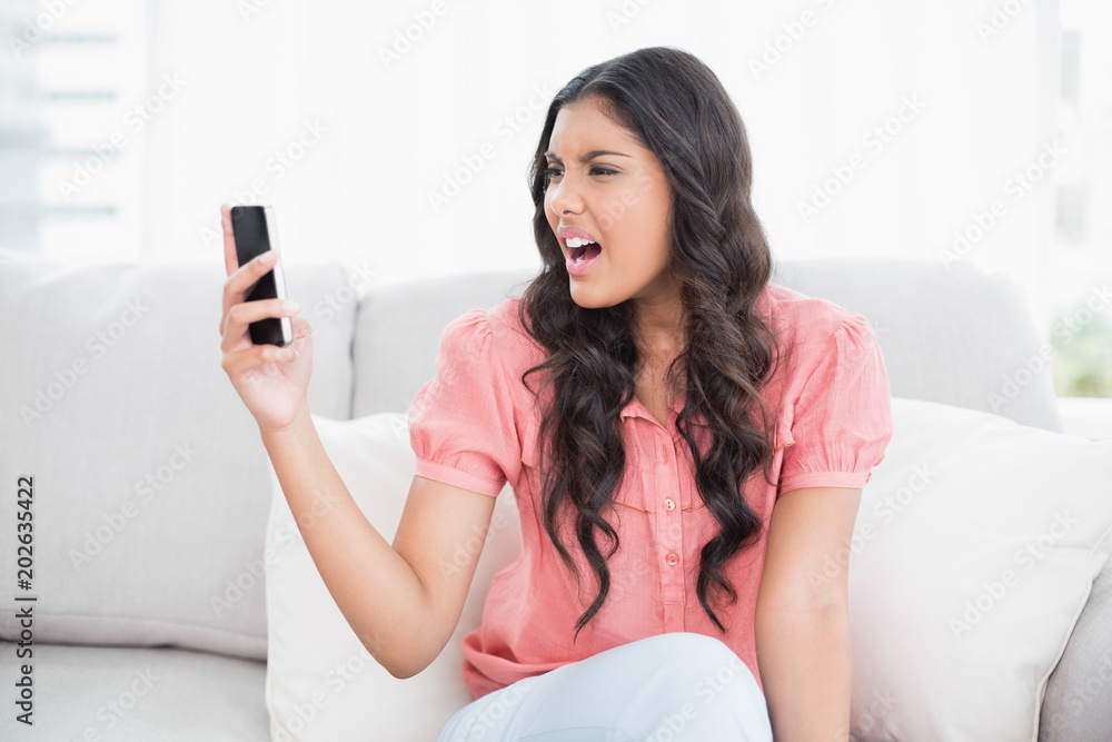 Angry cute brunette sitting on couch holding smartphone Angry cute brunette sitting on couch holding smartphone