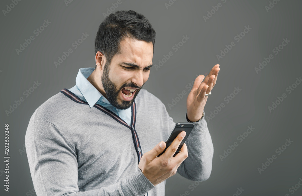 Bearded handsome man with angry face expression holding smartphone in hand on grey isolated background Bearded handsome man with angry face expression holding smartphone in hand on grey isolated background