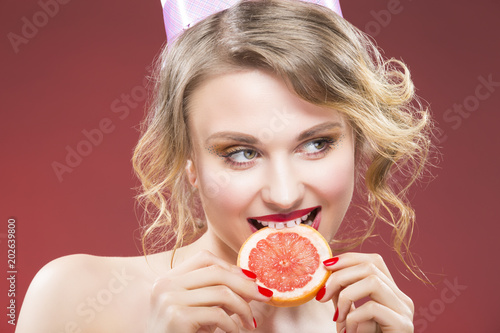 Fruit Ideas. Shy and Sexy Caucasian Blond Girl Biting Grapefruit Slice in Front. Against Red Background.