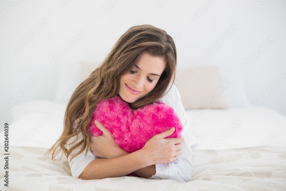 Happy casual brown haired woman in white pajamas hugging a heart shaped pillow Happy casual brown haired woman in white pajamas hugging a heart shaped pillow