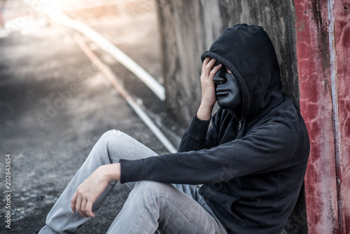 Mystery man with black mask and hoody jacket in headache gesture feeling stressed and depressed while sitting near grunge red concrete wall in abandoned building. major depressive disorder concept