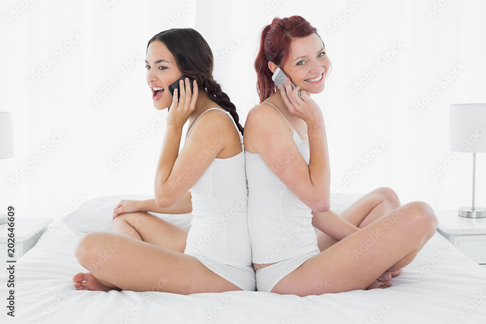 Friends using mobile phone while sitting back-to-back on bed Friends using mobile phone while sitting back-to-back on bed