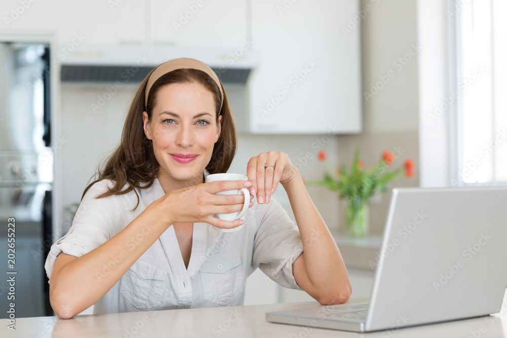 Smiling woman with coffee cup and laptop in kitchen Smiling woman with coffee cup and laptop in kitchen