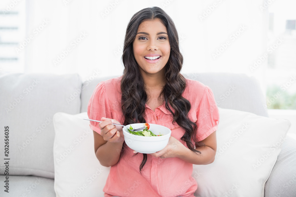 Happy cute brunette sitting on couch holding salad bowl Happy cute brunette sitting on couch holding salad bowl