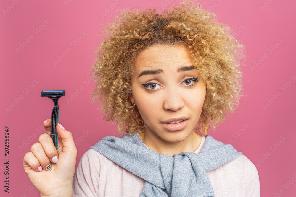Girl is wondering. She has a razer is her hand. Young woman is looking straight to the camera. She keeps her one brow up on the forehead. Isolated on pink background. Girl is wondering. She has a razer is her hand. Young woman is looking straight to the camera. She keeps her one brow up on the forehead. Isolated on pink background.