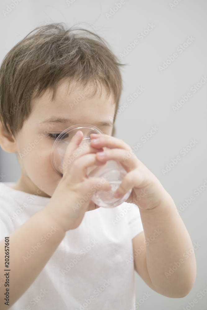 Handsome serious sweet little boy child with brown hair hazel eyes looking away sitting at table drinking water from small glass holding by hands indoor on blur grey background closeup, horizontal Handsome serious sweet little boy child with brown hair hazel eyes looking away sitting at table drinking water from small glass holding by hands indoor on blur grey background closeup, horizontal