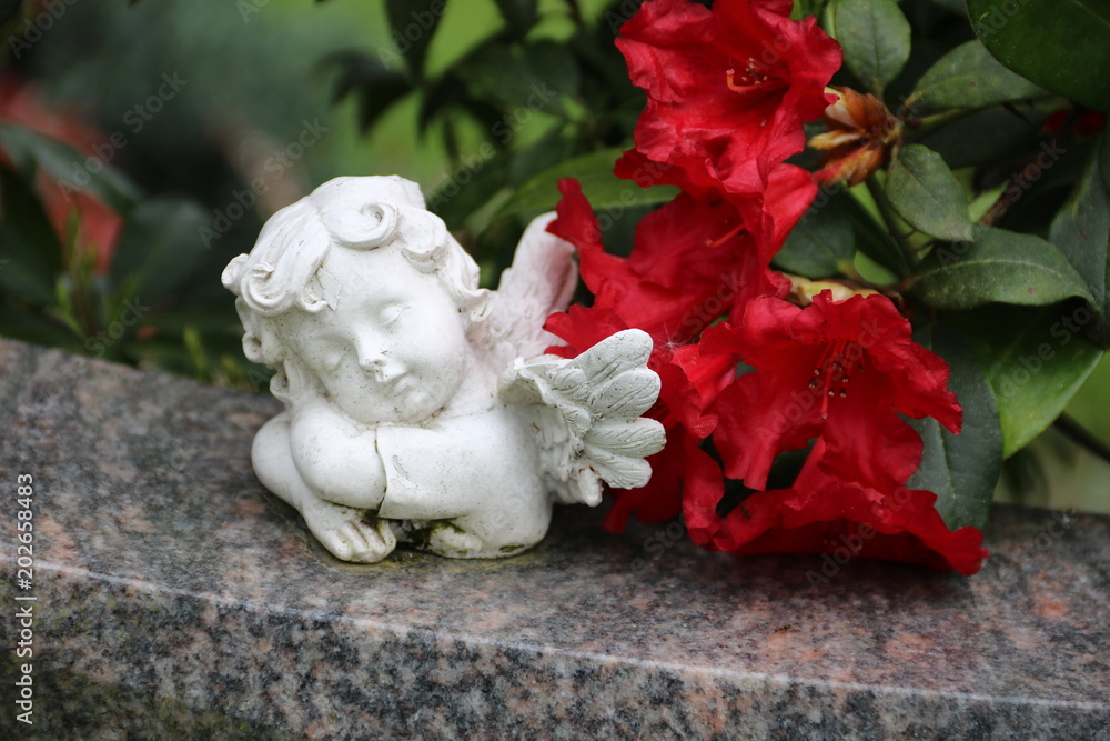 Kleiner Grabengel auf Grabstein neben Blüte einer Azalee in rot Kleiner Grabengel auf Grabstein neben Blüte einer Azalee in rot
