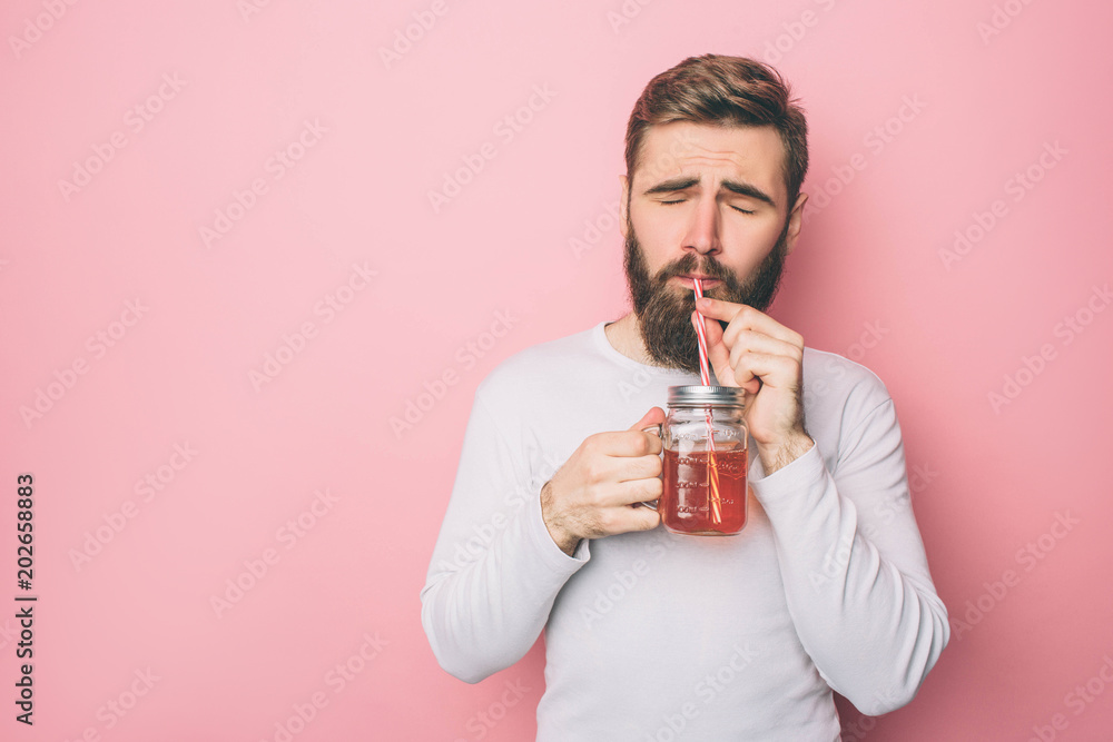 Bearded guy in a white sweater is drinking some tea from the cup though the straw. He is enjoying the moment. Isolated on pink background. Bearded guy in a white sweater is drinking some tea from the cup though the straw. He is enjoying the moment. Isolated on pink background.