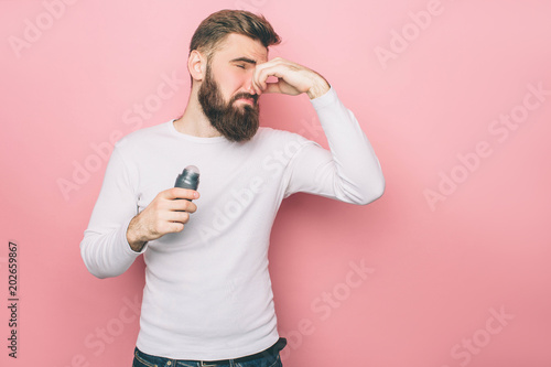 There is a bad smell in the room that goes from the armpits. Guy is closing his nose. It is disgusting. Guy is holding deodorant in his hand. Isolated on pink background.