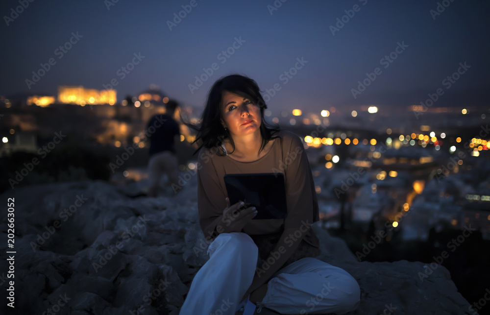 Beautiful woman with a tablet computer is sitting on a rock above the city lights of athens