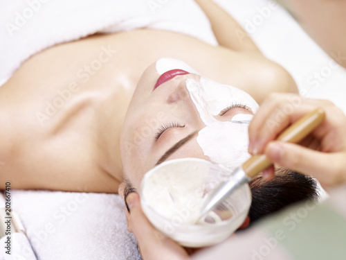 asian beautician applying facial mask on face of young woman