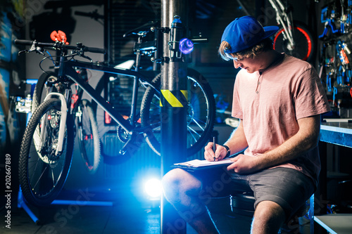 Young handsome stylish man in a cap snapback and with a tattoo small business owner selling a bicycle, the workshop sits on the background of the store in the hands of a notebook to write notes