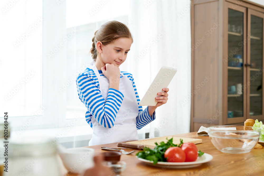 Curious little girl wrapped up in reading recipe with help of digital tablet while standing at wooden kitchen table in order to make surprise for Mothers Day, portrait shot Curious little girl wrapped up in reading recipe with help of digital tablet while standing at wooden kitchen table in order to make surprise for Mothers Day, portrait shot