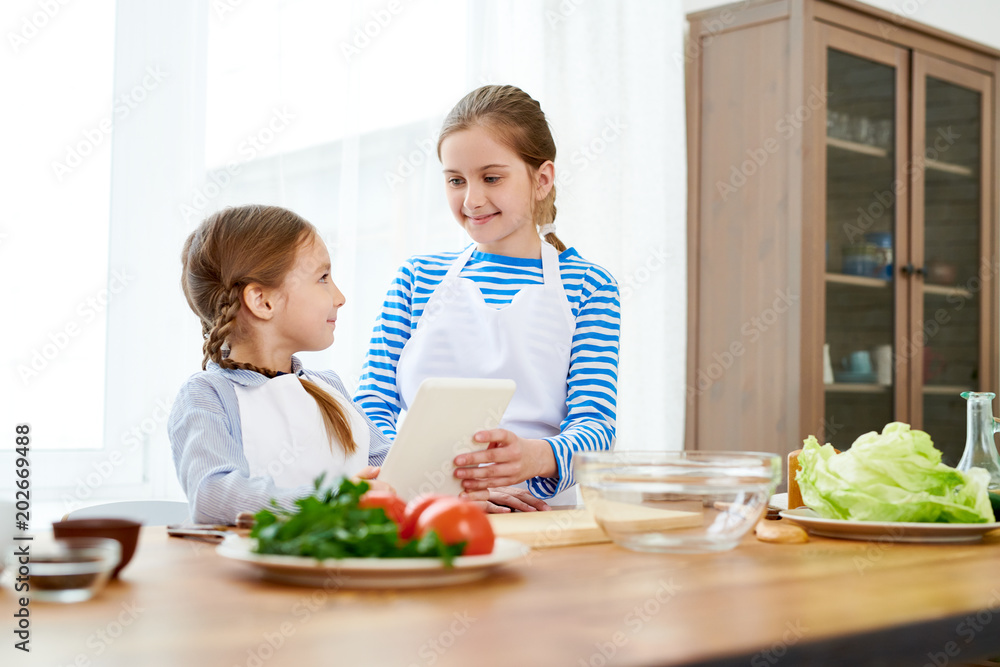 Cheerful little sisters wearing aprons looking at each other with wide smiles while preparing appetizing starter for their parents, spacious kitchen interior on background Cheerful little sisters wearing aprons looking at each other with wide smiles while preparing appetizing starter for their parents, spacious kitchen interior on background