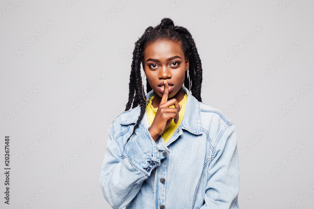 Beautiful Afro-American girl in casual clothes is showing silence sign finger on lips and looking at camera, isolated on gray Beautiful Afro-American girl in casual clothes is showing silence sign finger on lips and looking at camera, isolated on gray