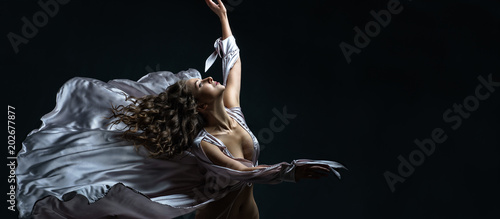 Beautiful brunette girl with curly hair in the darkness and light in sexy silver satin flying dress awesome poses in dance. Portrait art photo shooting in studio with female model