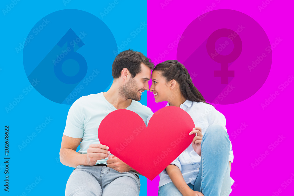 Cute couple sitting holding red heart against pink and blue Cute couple sitting holding red heart against pink and blue