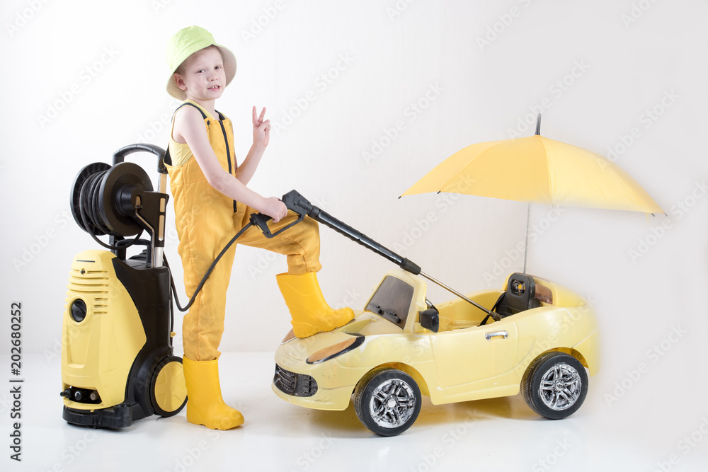 Happy child in yellow overalls and boots with high pressure washer serves client boy on toy car under umbrella. Concept of family business. White background Happy child in yellow overalls and boots with high pressure washer serves client boy on toy car under umbrella. Concept of family business. White background