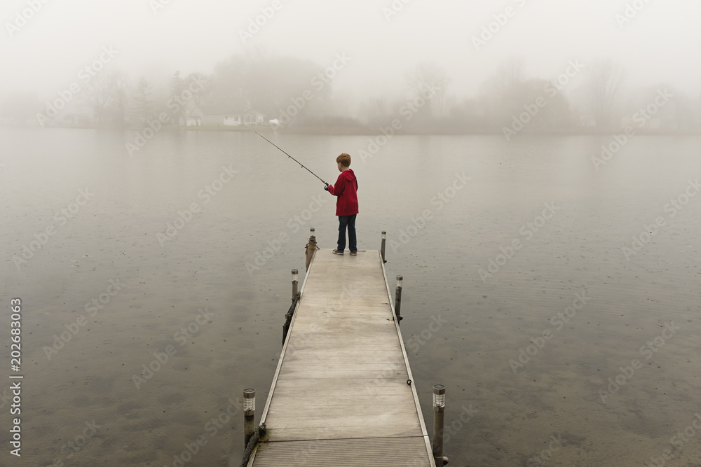 Young red hair boy fishing off a dock on a lake with fog