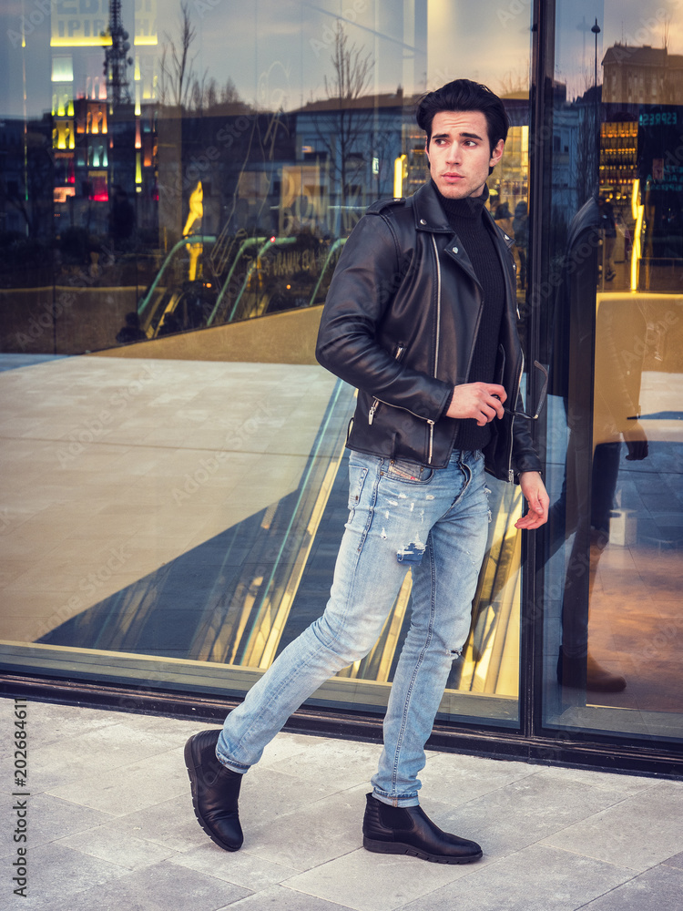 Obraz premium One handsome young man in urban setting in modern city, walking, wearing black leather jacket and jeans