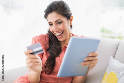 Excited brunette sitting on her sofa using tablet to shop online
