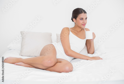 Peaceful young brown haired model in white pajamas drinking coffee