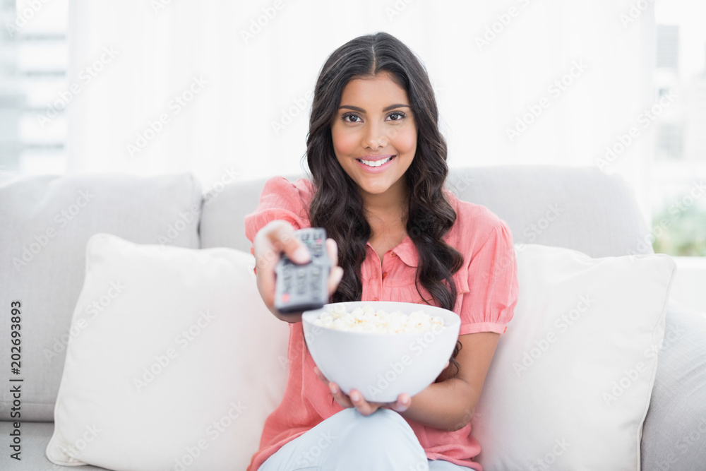 Smiling cute brunette sitting on couch holding popcorn bowl Smiling cute brunette sitting on couch holding popcorn bowl