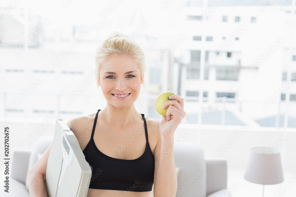 Smiling fit woman holding scale and apple in fitness studio Smiling fit woman holding scale and apple in fitness studio