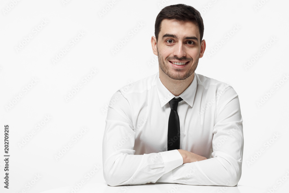 Indoor picture of handsome Caucasian male dressed in white shirt and necktie pictured isolated on white background resting arms on white table feeling optimistic and ready for business communication Indoor picture of handsome Caucasian male dressed in white shirt and necktie pictured isolated on white background resting arms on white table feeling optimistic and ready for business communication