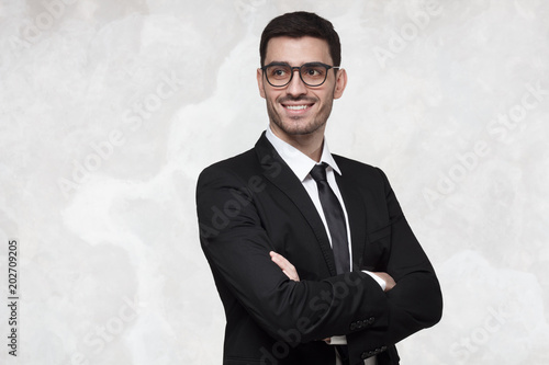 Half-length portrait of handsome European guy or businessman isolated on bright background wearing formal suit and glasses, having arms crossed, showing happy confident smile, feeling relaxed photo