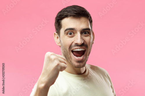 Horizontal portrait of young Caucasian guy pictured isolated on pink background wearing casual T-shirt smiling and laughing in excitement while supporting team or celebrating victory in competition