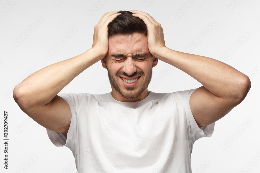 Closeup headshot of young European Caucasian man isolated on grey background dressed in white T-shirt pressing firmly hands to temples experiencing bad headache, having shut eyes because of pain Closeup headshot of young European Caucasian man isolated on grey background dressed in white T-shirt pressing firmly hands to temples experiencing bad headache, having shut eyes because of pain