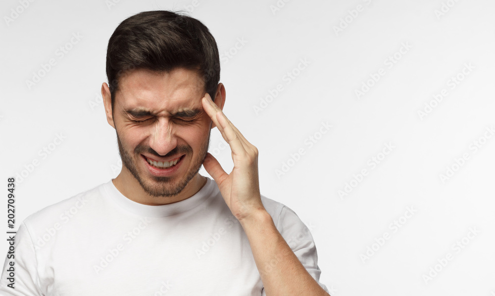 Indoor closeup of young European Caucasian man isolated on gray background in white casual T-shirt touching temple with fingers as if suffering from severe migraine, feeling helpless and sick Indoor closeup of young European Caucasian man isolated on gray background in white casual T-shirt touching temple with fingers as if suffering from severe migraine, feeling helpless and sick