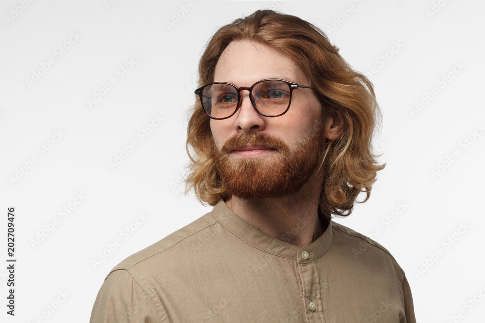 Horizontal closeup shot of young Scandinavian guy with moustache and beard pictured isolated on gray background smiling and looking aside as if looking forward to something new and positive in life Horizontal closeup shot of young Scandinavian guy with moustache and beard pictured isolated on gray background smiling and looking aside as if looking forward to something new and positive in life
