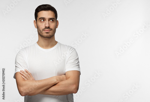 Indoor photo of young European Caucasian man isolated on gray background dressed in white casual T-shirt standing with arms crossed looking upward as if thinking, dreaming or looking for idea