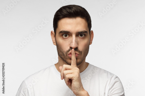 Horizontal photo of handsome European man isolated on gray background dressed in white casual T-shirt pressing finger to lips as if asking to keep silence, not willing anyone to know secret