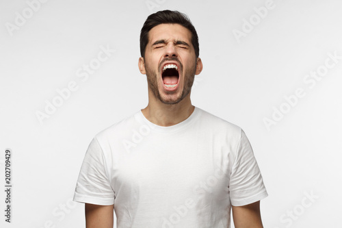 Picture of young European Caucasian man isolated on grey background wearing white T-shirt standing with eyes shut and mouth open as if screaming in despair or being extremely exhausted photo