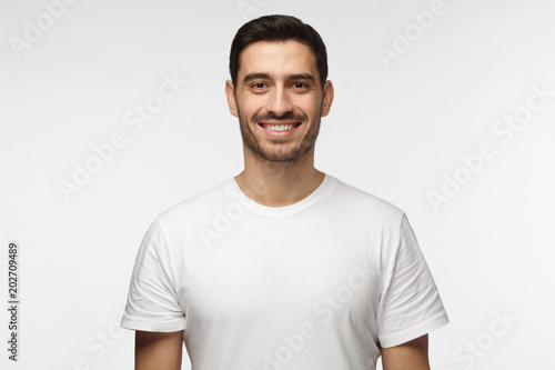 Horizontal shot of young positive European guy isolated on gray background standing in white casual T-shirt looking straight at camera with happy smile, feeling relaxed, confident and satisfied
