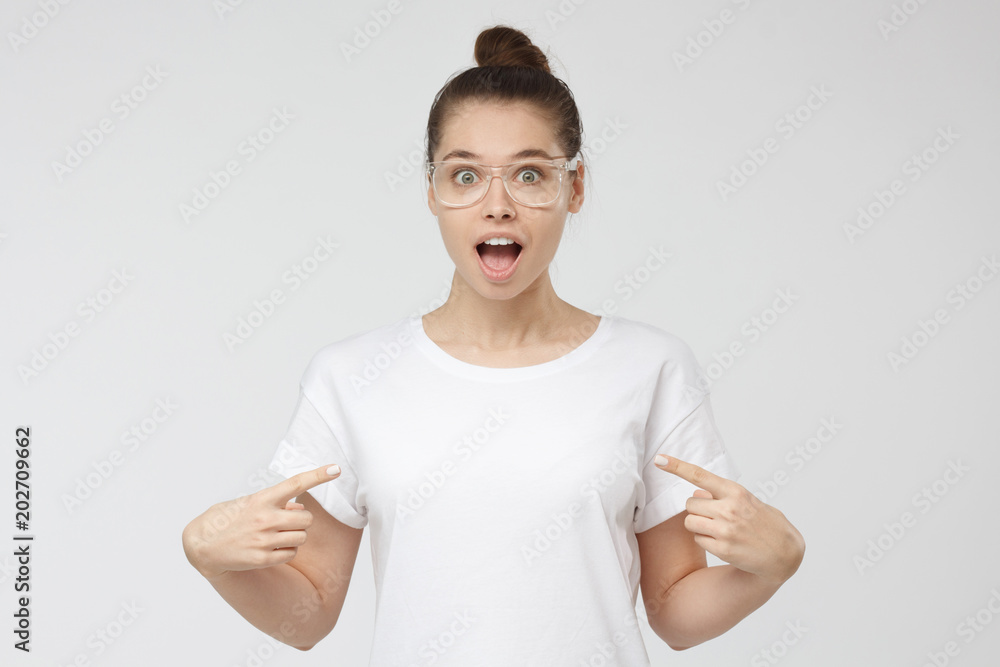 Horizontal portrait of young beautiful European female isolated on gray background wearing transparent glasses and white blank T-shirt pointing to it with surprised face, copyspace for advertising Horizontal portrait of young beautiful European female isolated on gray background wearing transparent glasses and white blank T-shirt pointing to it with surprised face, copyspace for advertising