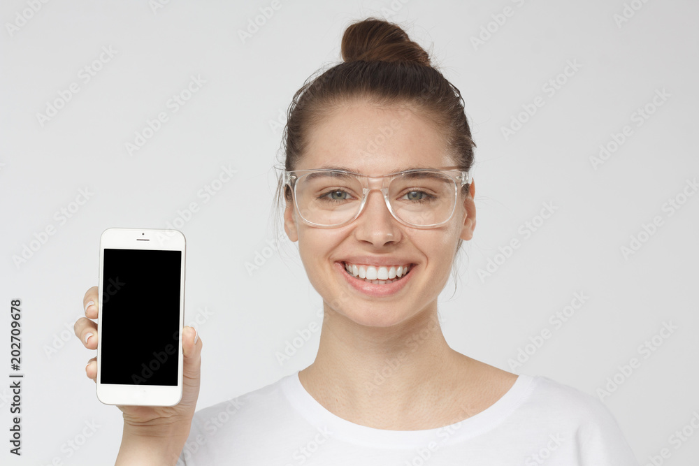 Indoor headshot of young beautiful European lady in transparent glasses without makeup isolated on gray background smiling happily while showing smartphone with blank screen, copyspace for advertising Indoor headshot of young beautiful European lady in transparent glasses without makeup isolated on gray background smiling happily while showing smartphone with blank screen, copyspace for advertising