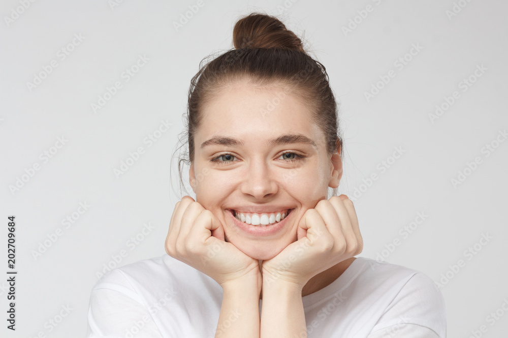 Closeup portrait of young pretty teenage girl isolated on gray background pressing fists to chin and smiling positively at camera looking attentively at person she is listening with love and care Closeup portrait of young pretty teenage girl isolated on gray background pressing fists to chin and smiling positively at camera looking attentively at person she is listening with love and care
