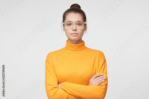 Closeup photo of young beautiful woman pictured isolated on grey background in mustard casual sweater and round glasses, standing with arms crossed and serious concentrated face at her partner photo