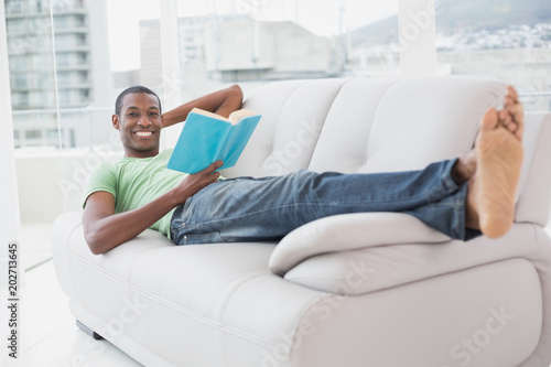 Full length portrait of smiling Afro man reading a book on sofa